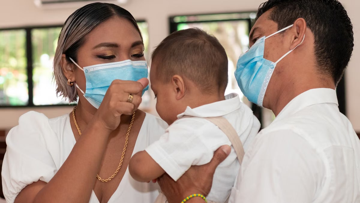 A family with a baby wearing face masks, indoors, during a social event.